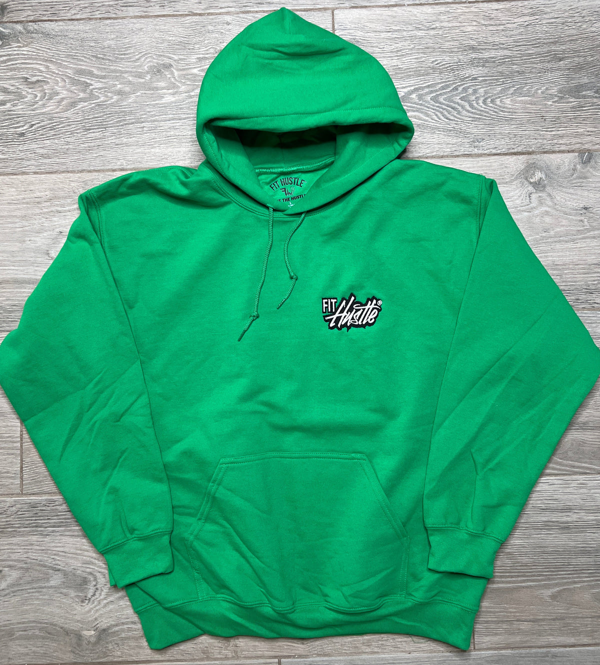 A bright green pullover hoodie with a small white and black 'Fit Hustle' logo on the left chest, laid flat on a gray wood plank floor.
