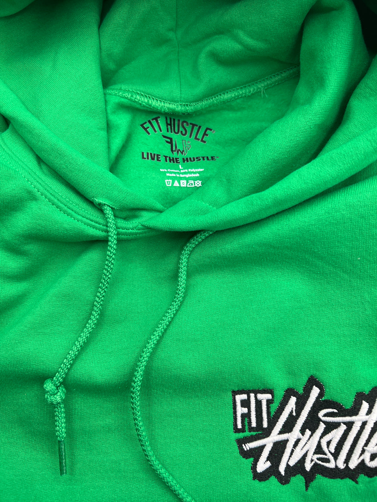 Close-up of a bright green hoodie with matching drawstrings and a black-and-white 'FIT Hustle' logo printed on the lower right chest. The inner neck label also reads 'FIT HUSTLE' and 'LIVE THE HUSTLE'.